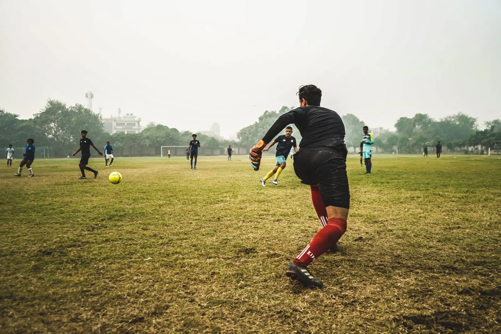 Boys playing football on a wide open ground in the evening, one running at the ball in the front.