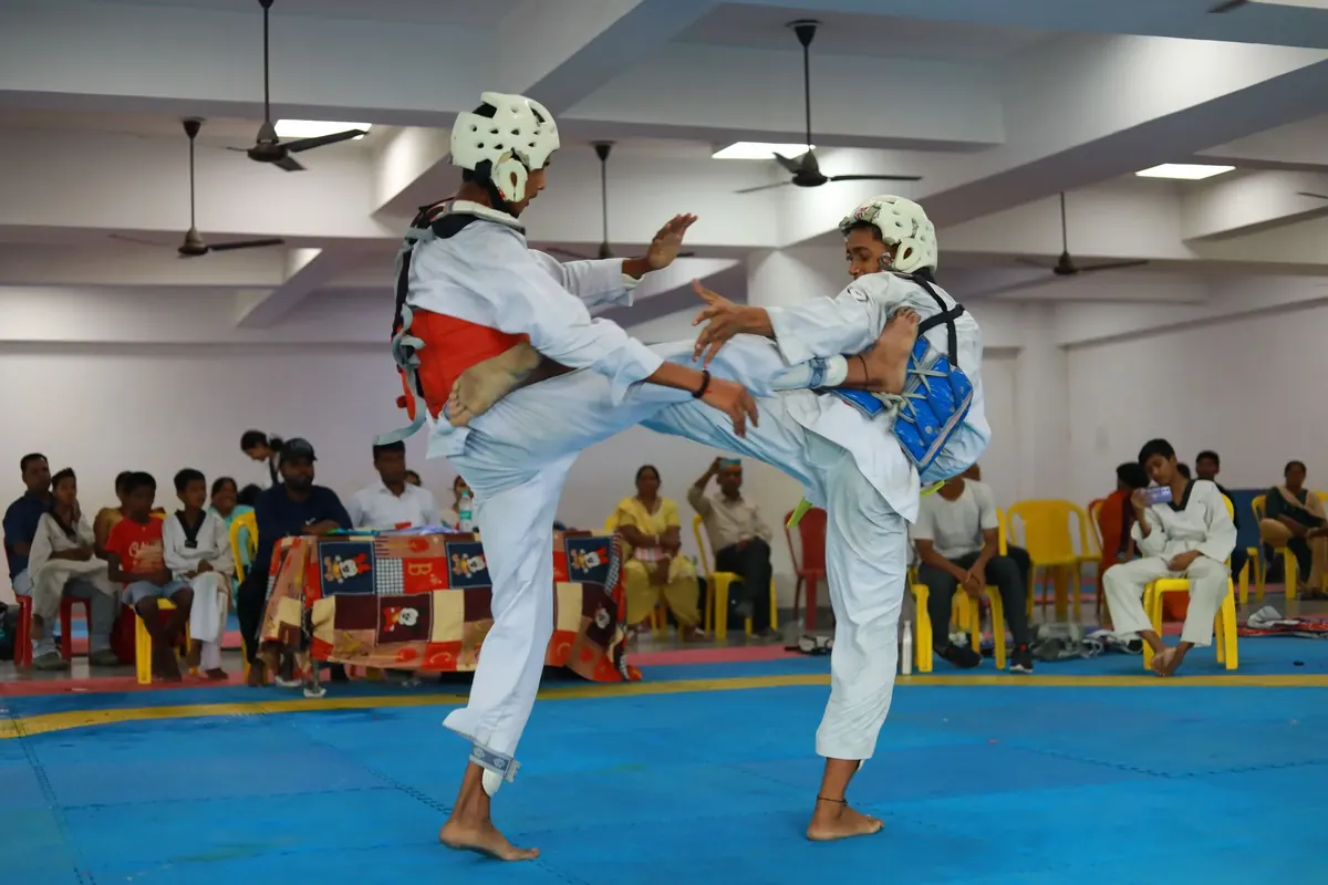 Two young taekwondo players in headgear and chest guards trade kicks in an indoor arena while families watch.