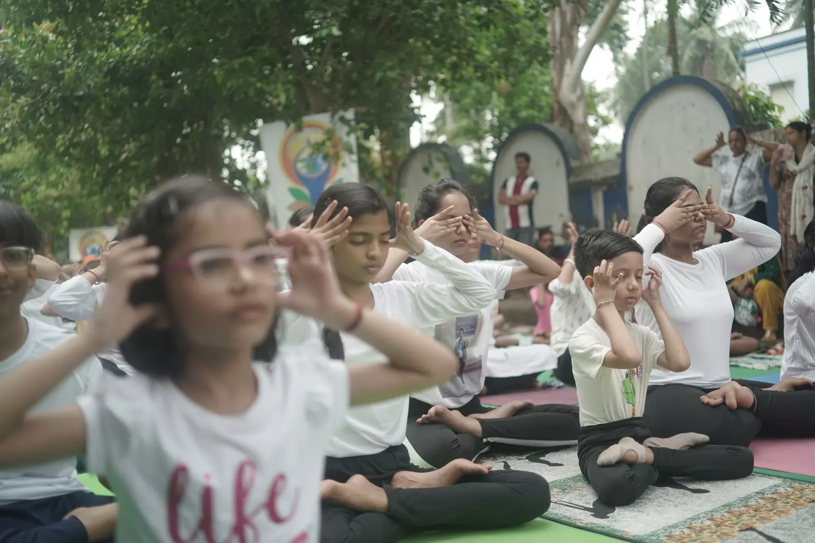 Children in white T-shirts sit cross-legged on mats for an outdoor yoga class under the trees.